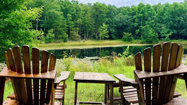 Two wooden Adirondack chairs and a picnic table facing a lake surrounded by dense forest.