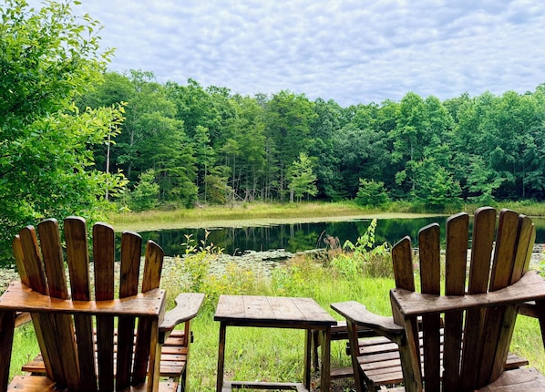 Two wooden Adirondack chairs and a picnic table facing a lake surrounded by dense forest.