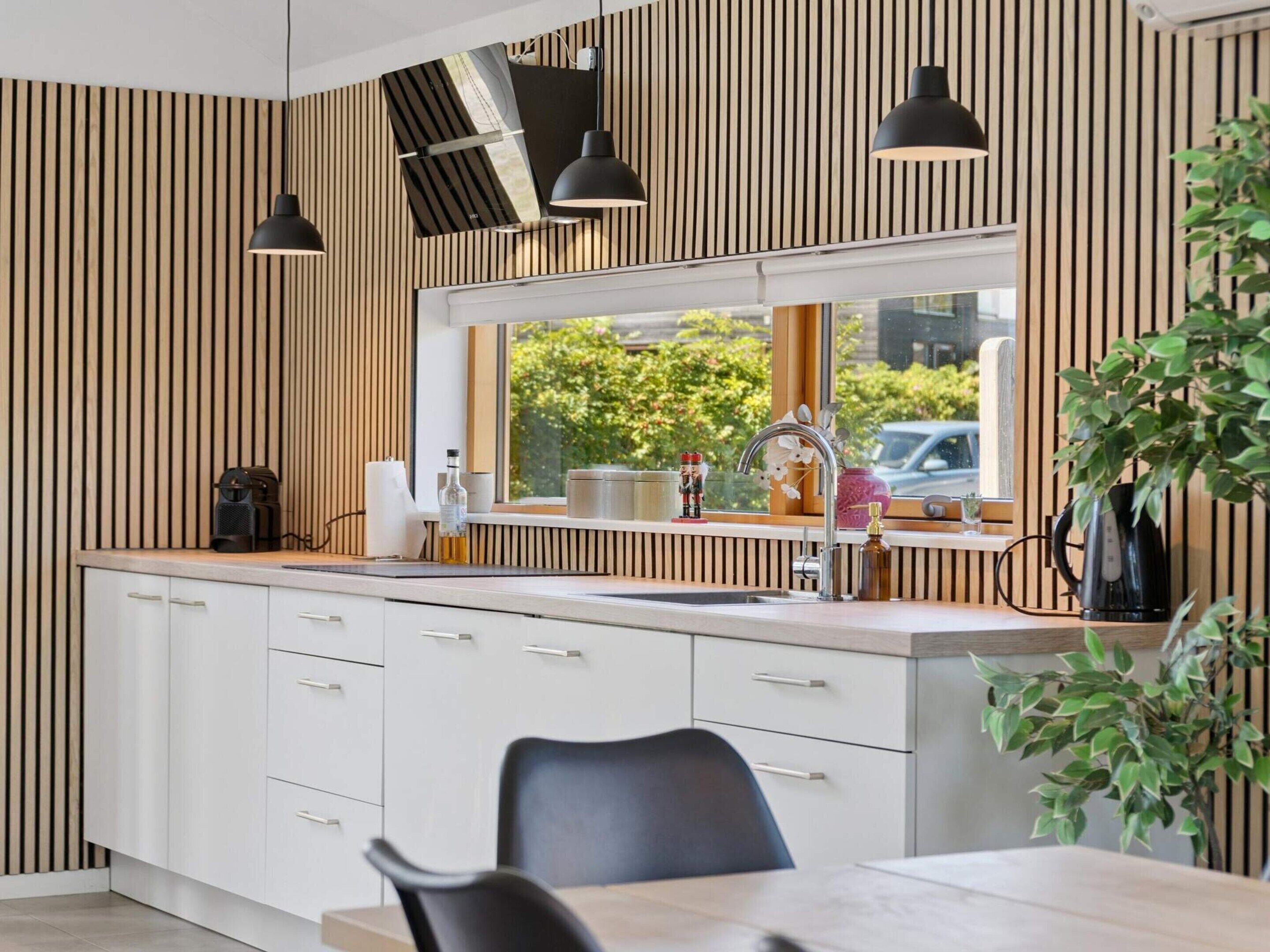 A modern kitchen with a window, wooden panel wall, and black pendant lights.