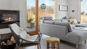 A modern living room with a fireplace, grey sofa, wicker chair, and a small stool. - 4 Star Holiday Home in Stubbekobing (Stubbekobing)