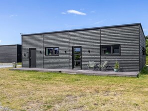 Een modern, gelijkvloers gebouw met een plat dak, grote ramen en een klein terras met twee stoelen.