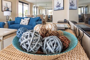 Une pièce comprenant une table en osier sur laquelle sont posés plusieurs pelotes de laine, un canapé bleu agrémenté de coussins à motifs et des œuvres d’art encadrées accrochées aux murs.