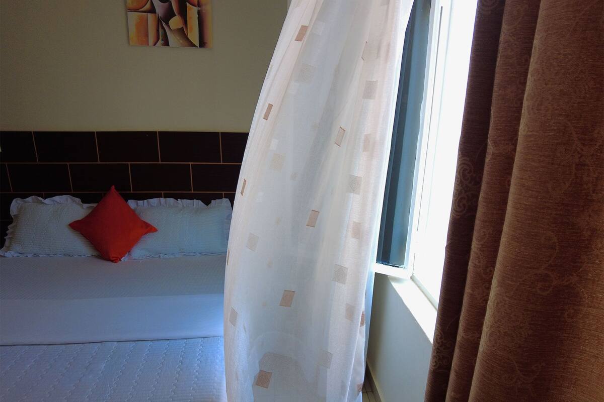 A hotel room with a bed, a headboard, a window with curtains, and a wall with a painting.