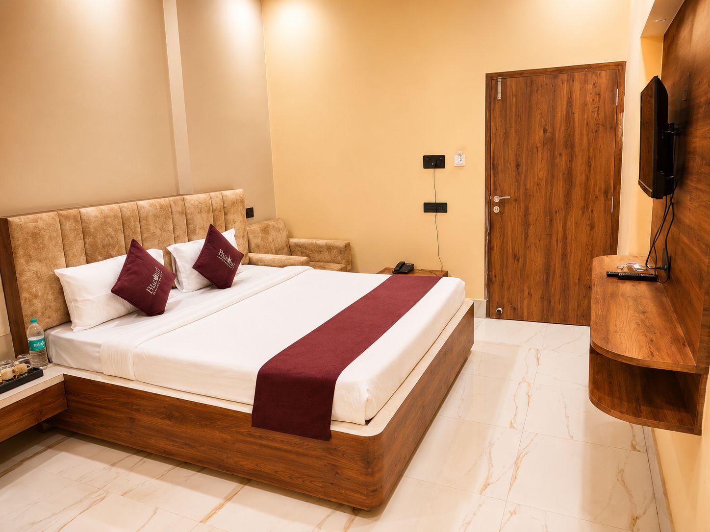 Habitación de hotel con estructura de cama de madera, televisor de pantalla plana en la pared, mesita de noche con teléfono y puerta de madera.