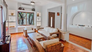 A living room with a sofa, coffee table, and TV. There is a window with a view of trees, a door, and a wall-mounted shelf with decorative items. - Beautiful 4-bedroom Villa with Private Pool close to Hotel Zone (Cancún)