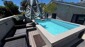 Una piscina en la azotea con terraza de madera, sillas de descanso y un área de estar cubierta.