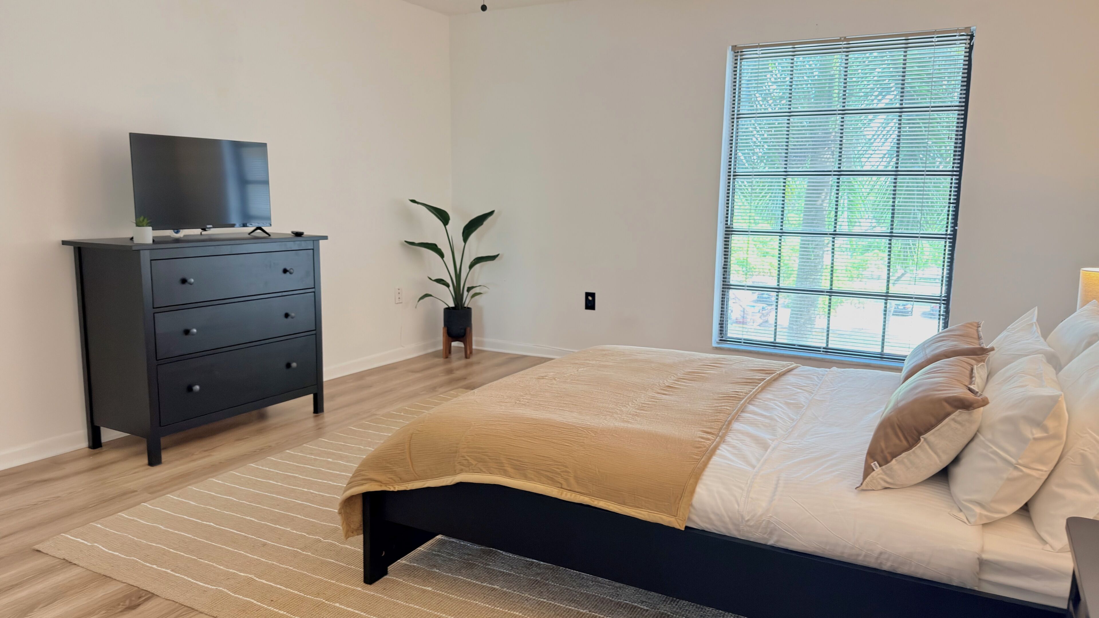 Un dormitorio con cama, cómoda, planta en maceta y ventana con persianas.