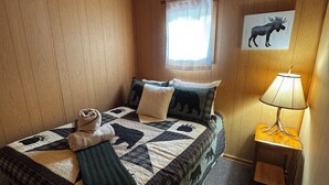 Una habitación con paneles de madera, una cama, una ventana con cortina, una lámpara y un cuadro de un alce en la pared.