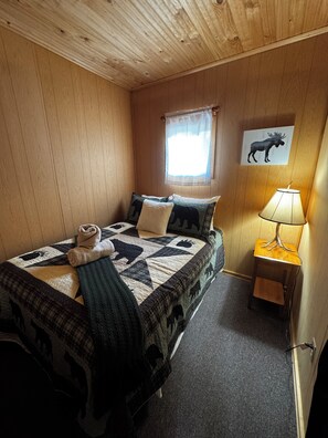 A wooden-paneled room with a bed, a window with a curtain, a lamp, and a moose picture on the wall.