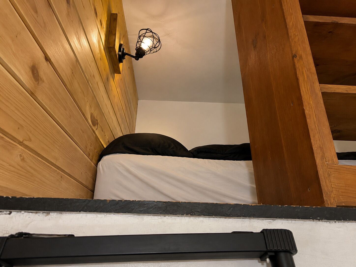 Un llit superposat de fusta amb capçalera, una làmpada de paret i una prestatgeria negra.