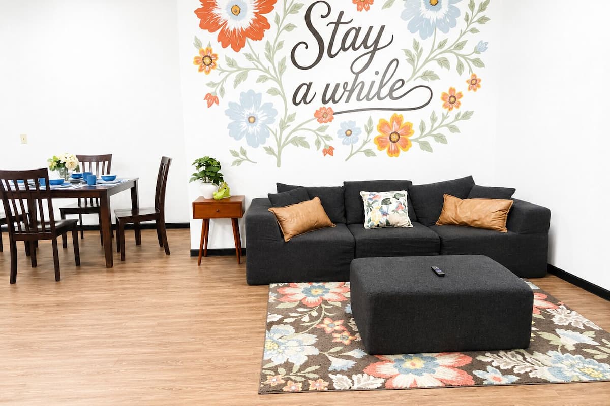A living room with a black sofa, a matching ottoman, a floral rug, a dining table with chairs, and a wall with a floral decal and the text 'Stay a while'.