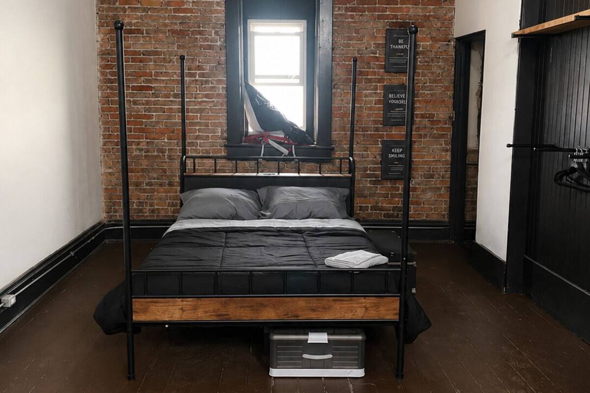 A single bed with a wooden frame and black bedding, a brick wall, a window with a view, and a black door.