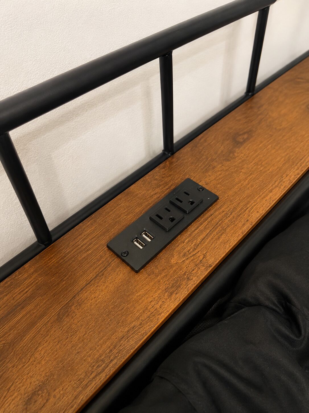 A wooden bedside table with a black power strip and a black metal headboard.