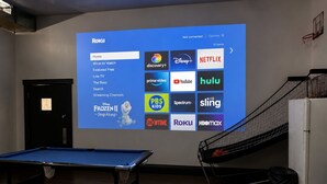 A room with a pool table, a projector displaying a Roku interface, and a basketball hoop.