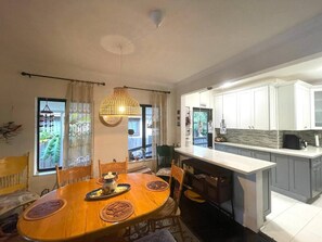Zona pranzo con tavolo e sedie in legno, una lampada a sospensione e vista sulla cucina con mobili bianchi.