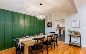 Ein Essbereich mit Holztisch, Stühlen, einer Chandelüster und einer Küche im Hintergrund.