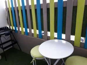 Ein kleiner Sitzbereich im Freien mit einem weißen Tisch, zwei grünen Stühlen, einem Grill und einem bunten Zaun.