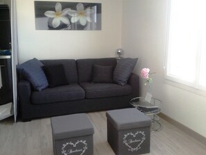 Ein modernes Wohnzimmer mit einem dunkelgrauen Sofa, einem Glastisch und zwei grauen Poufs mit Herzmustern.