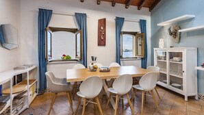 A dining area with a wooden table, white chairs, a cabinet with glass doors, and blue curtains. - La Casetta Azzurra (Aci Castello)