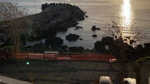 A coastal view with a rocky shoreline, a clear sky, and the sun setting or rising over the water. - La Casetta Azzurra (Aci Castello)