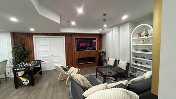 A modern living room with a foosball table, a sofa, a television displaying Netflix, and a fireplace.