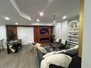 Uma sala de estar moderna com mesa de futebol, sofá, televisão exibindo Netflix e lareira.