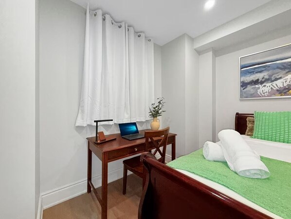 A hotel room with a wooden desk, a laptop, a green bedspread, and a painting on the wall.