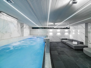 A modern indoor swimming pool with a tiled floor, a seating area with a sofa and a towel, and a ceiling with linear lighting fixtures. - Aman 15br2 Pools Park Lleras Events Ok (Medellín)