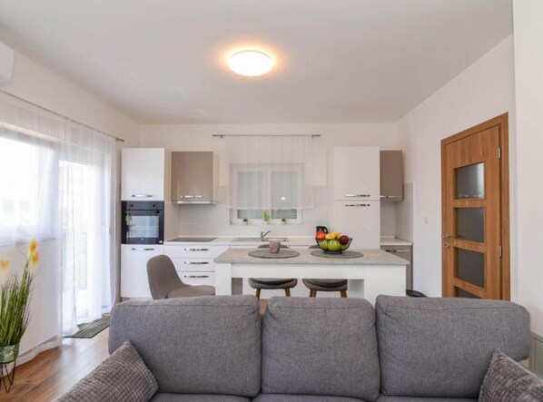A modern kitchen with white cabinets, a central island with a fruit bowl, and a dining area with a grey sofa. - One Bedroom Apartment with Terrace and Sea View (Sukosan)