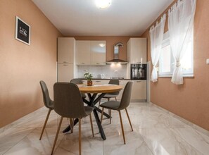 A dining area with a round table, four chairs, a kitchen in the background, and a window with curtains. - Two Bedroom Apartment with Terrace (Sukosan)