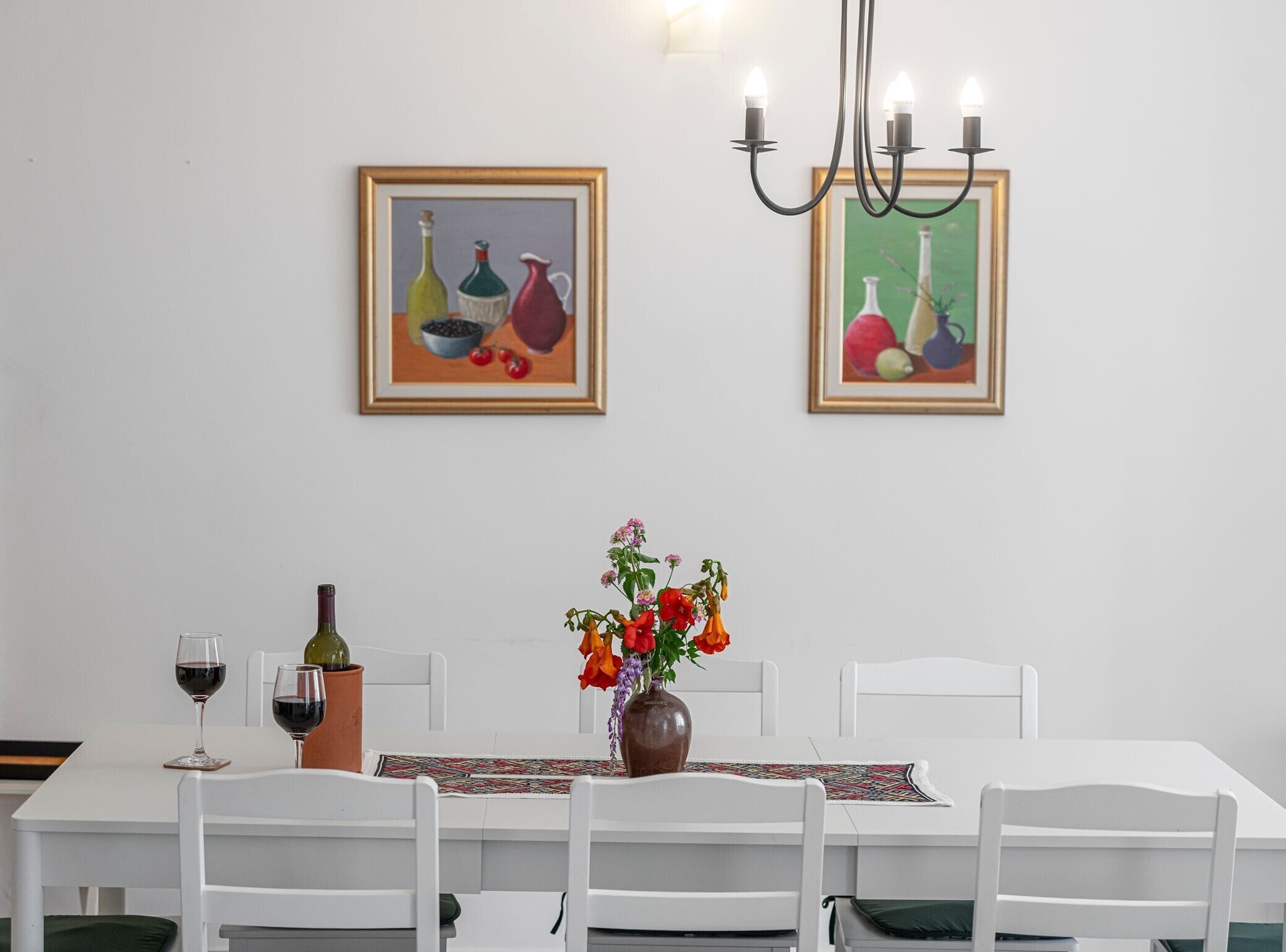 A dining area with a table set for two, two framed paintings, a vase with flowers, and a chandelier.