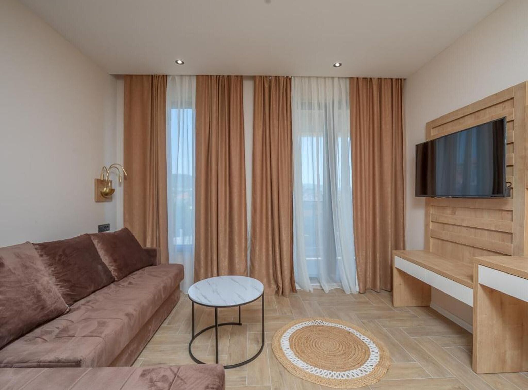 A modern living room with a sofa, a small round table, a TV mounted on a wooden cabinet, and large windows with sheer curtains.