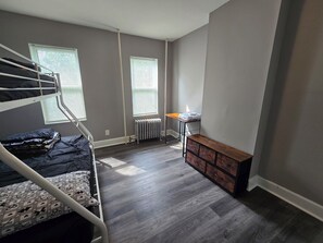 A room with a bunk bed, a radiator, a wooden bench, and a table with a lamp.