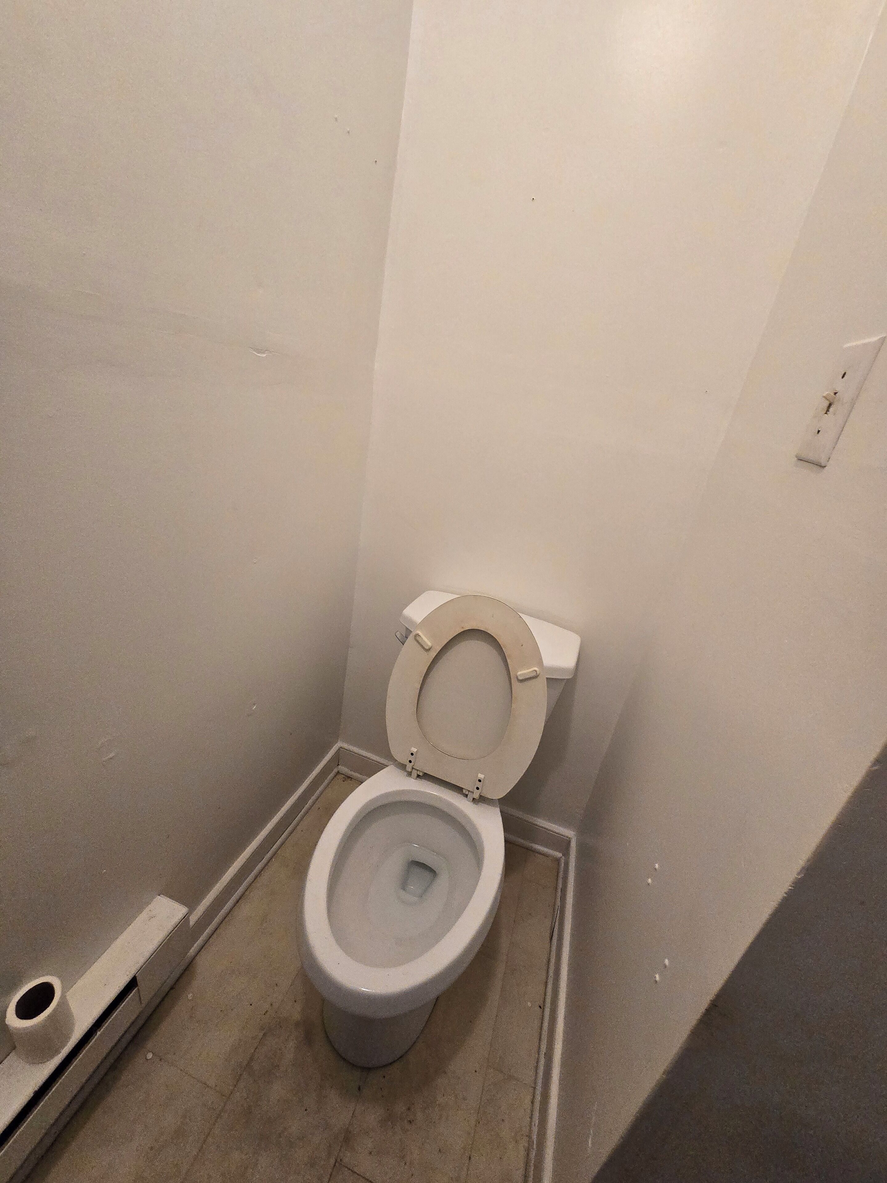 A small, narrow bathroom with a toilet, a wall-mounted light switch, and a floor drain.