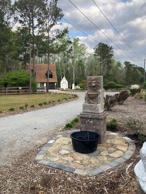 A stone lion statue with a water spout, surrounded by a stone and brick base, set in a landscaped area with a gravel path, trees, and a building in the background. - Cozy cabin in pleasant Little River perfect for a relaxing getaway. 6 min 2beach (Little River)