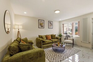 A living room with a sofa, armchair, and coffee table, a window with curtains, and a radiator. - New 4BR Home, Bedworth, Driveway Parking, Workspace, WiFi, Near M6 (Bedworth)