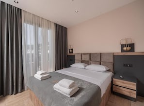 A modern hotel room with a large bed, a bedside table, a nightstand, and a window with curtains.