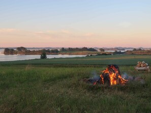 Ein Lagerfeuer auf einer grünen Wiese mit einem See und Bäumen im Hintergrund.