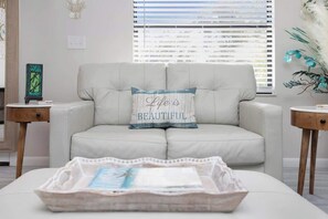 Ein helles Sofa mit einem Kissen mit der Aufschrift 'Life is Beautiful', ein Holznachttisch, eine Wanddekoration und ein Fenster mit Jalousien.