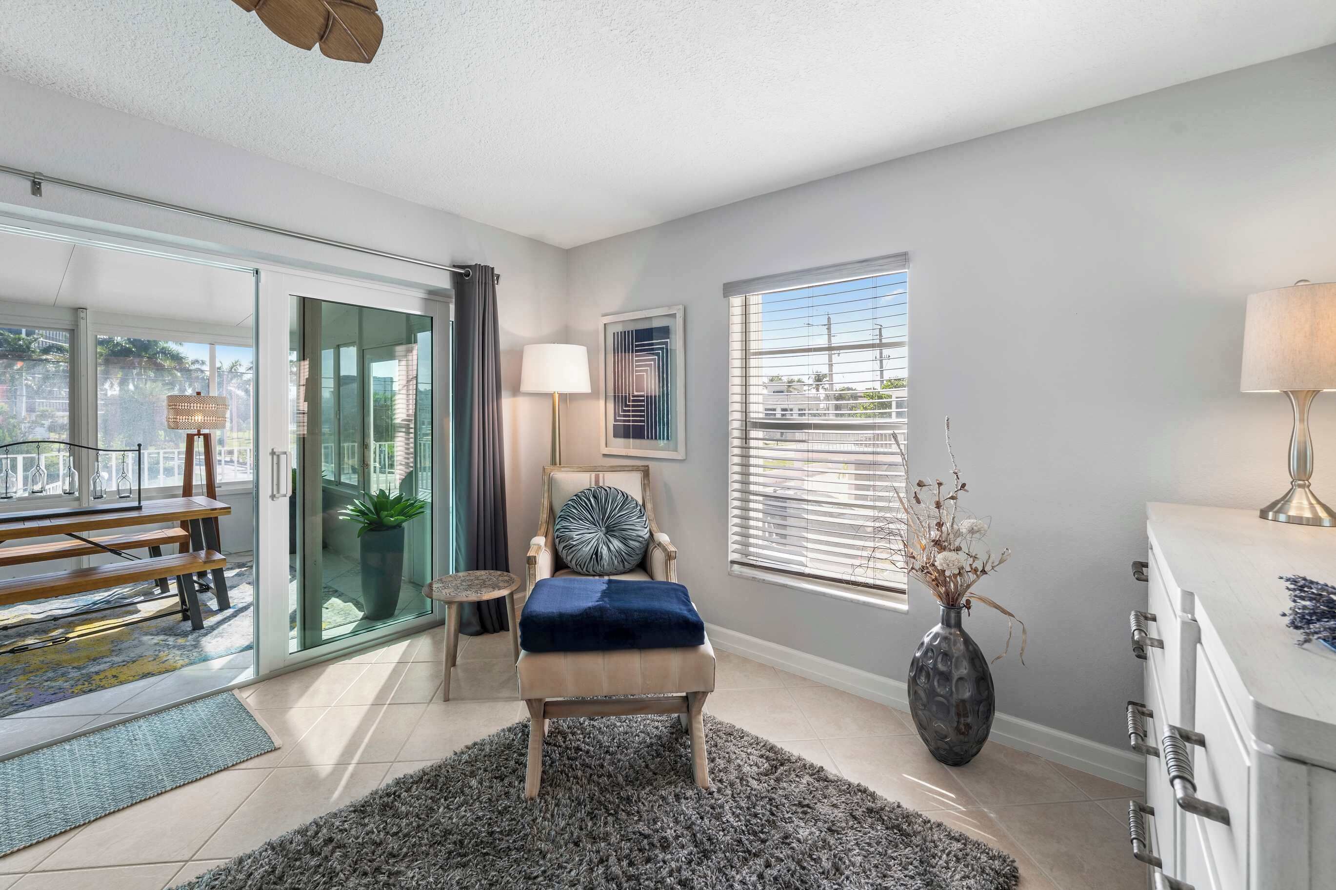A modern living room with a glass door leading to a balcony, a plush armchair, a side table with a lamp, and a vase with dried flowers.