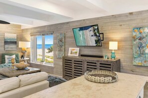 Ein modernes Wohnzimmer mit einem großen Flachbildfernseher, Holzpaneelen, einer gemütlichen Couch und Blick auf den Strand durch große Fenster.
