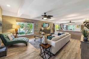 Ein geräumiger Wohnbereich mit einem grünen Sofa, einem weißen Couche und einem Holzboden. Deckenventilatoren, ein Fernseher und ein Essbereich sind sichtbar.