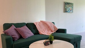 Un sofá verde con dos almohadas, una mesita redonda con una planta pequeña y una radio, y un cuadro en la pared.