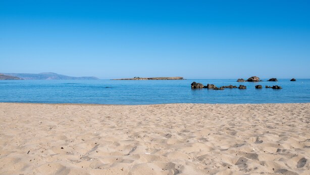 Een zandstrand met kalm water en verafgelegen rotsformaties.