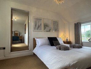 A bedroom with a bed, a nightstand, a window with curtains, and a hallway leading to another room. - Spacious 5 Bedroom Cottage (Gotham)