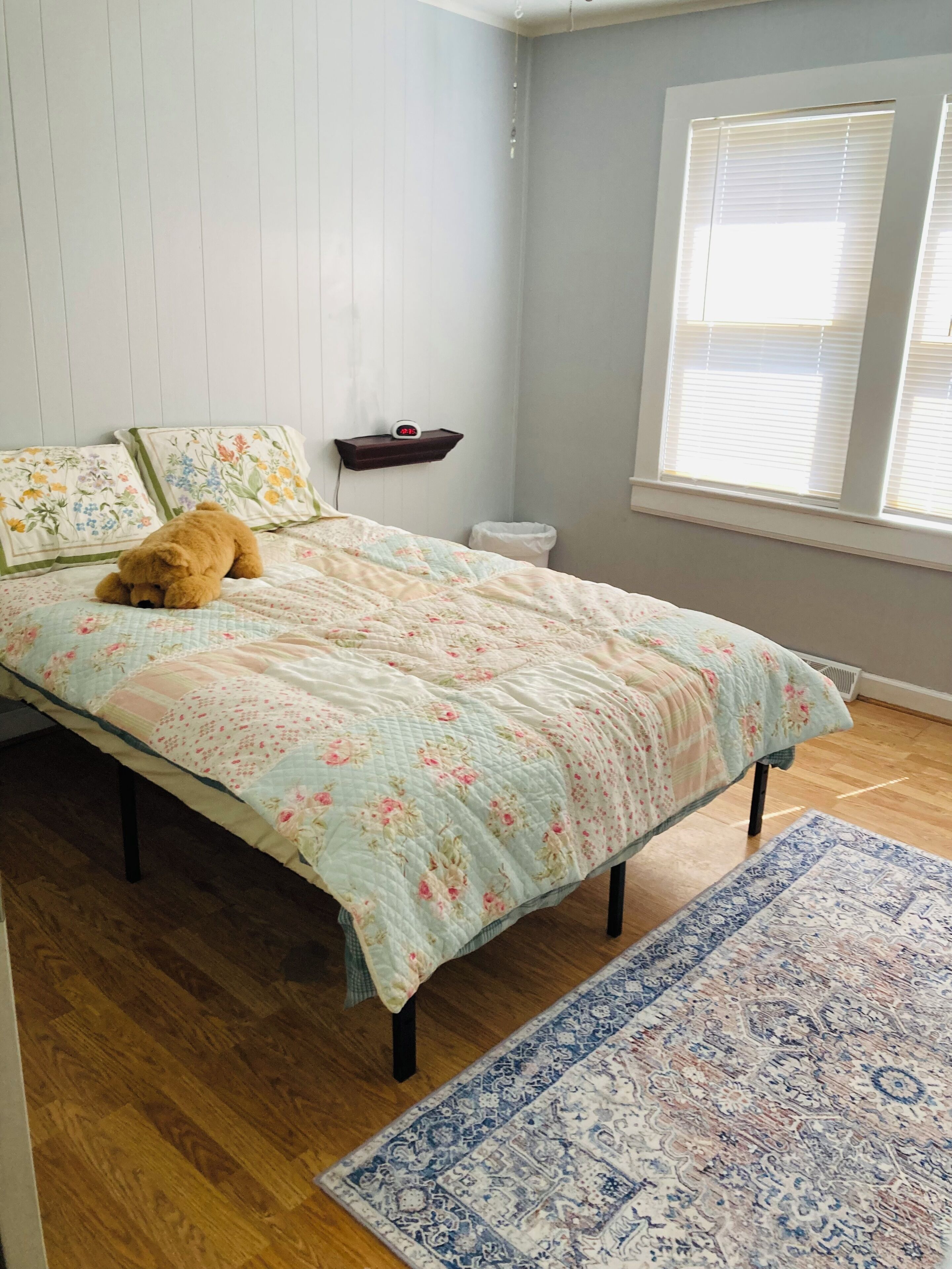 A bedroom with a bed, a nightstand, a clock, a window with blinds, and a wooden floor.