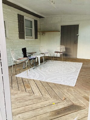 A covered patio area with a white patterned rug, wooden flooring, and white metal chairs.