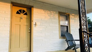 Une maison avec une porte beige, une petite fenêtre ornée d’un cadre décoratif et une véranda en bois avec une chaise.