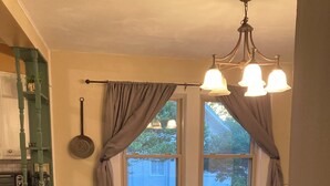 Un coin repas avec une table dressée pour deux personnes, un lustre et une vue sur l’extérieur par une fenêtre ornée de rideaux.