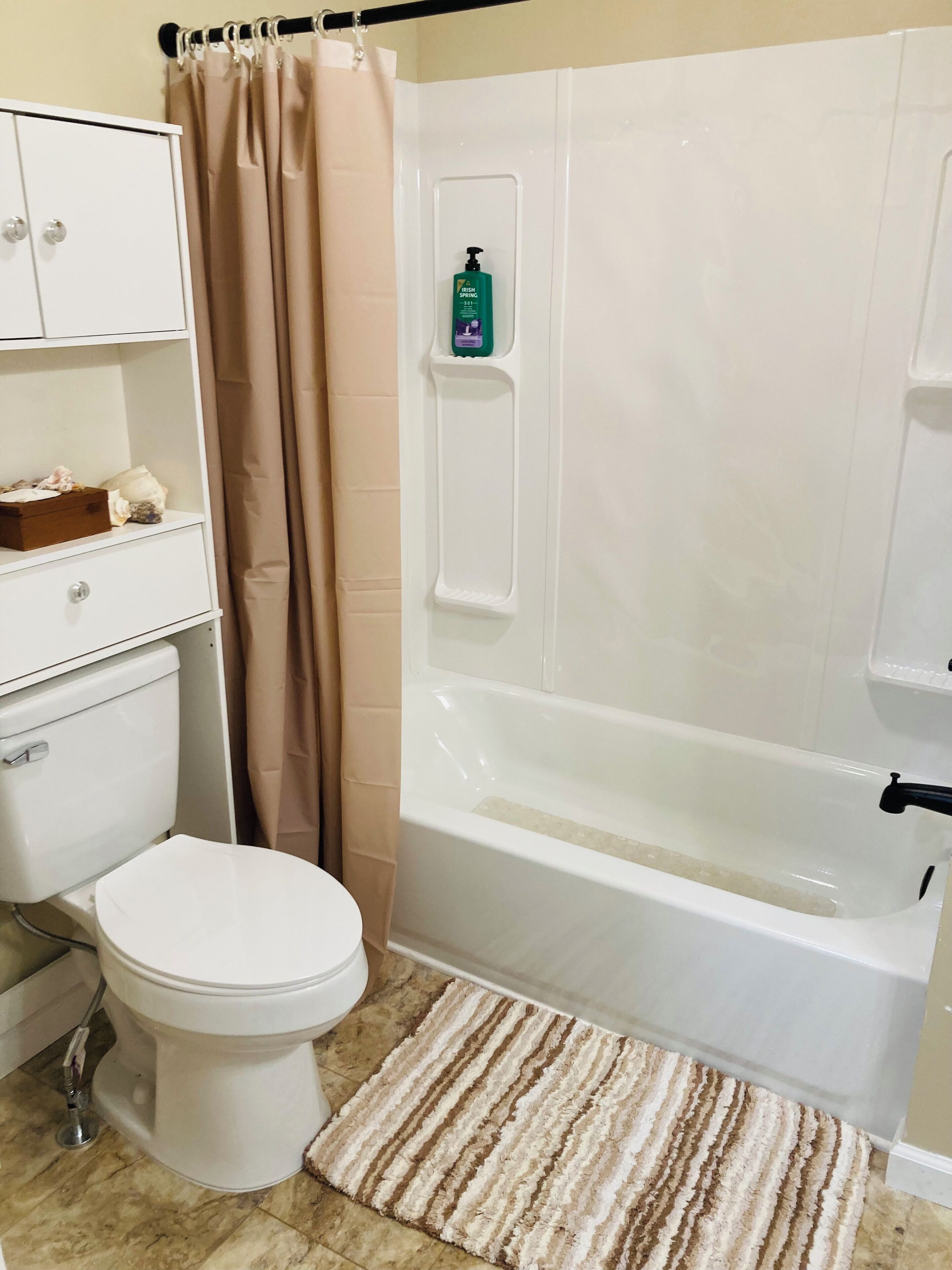 Une salle de bain comprenant des toilettes, une baignoire, un rideau de douche, une étagère avec des articles de toilette et un tapis à rayures.